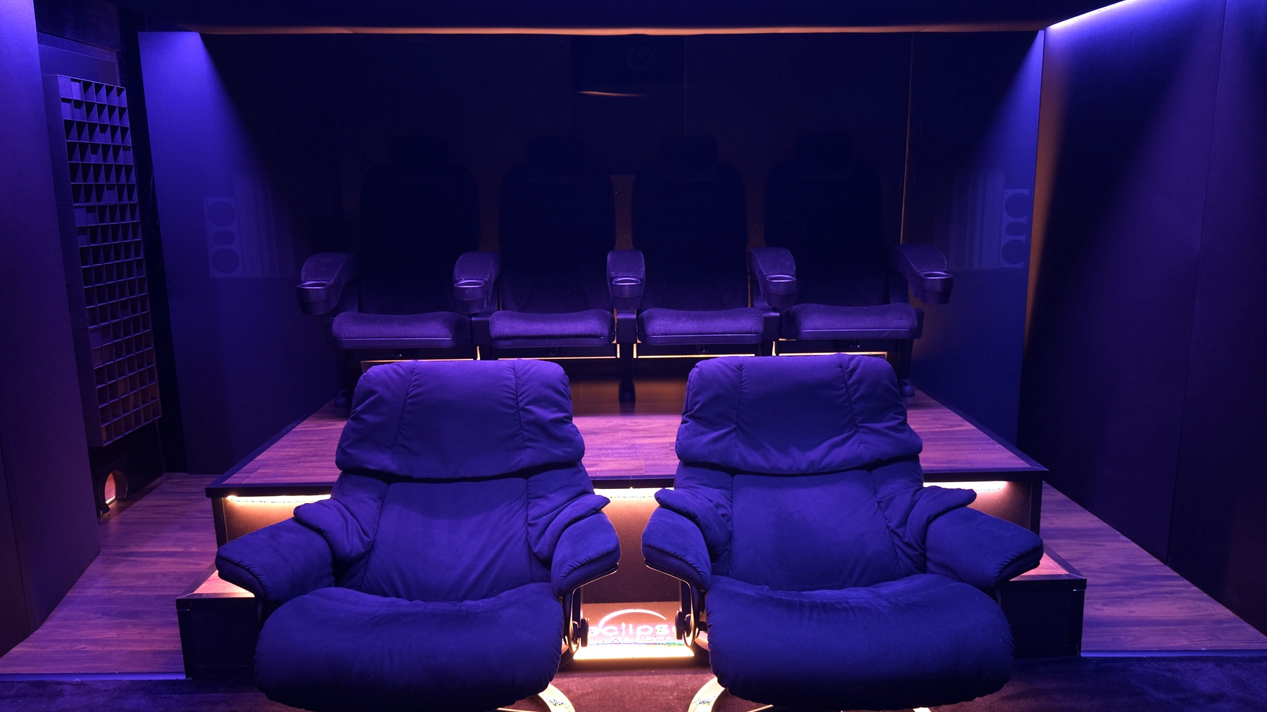 Blick vom Leinwandbereich auf Lounge-Sessel, Podest und hintere Sitzreihe im blau-violetten Heimkino